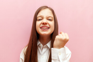 angry little teenage girl with dental braces threatens and shows fist on pink isolated background,...