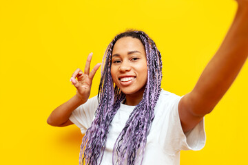 cheerful african american woman with colored dreadlocks making selfie and showing peace gesture on...