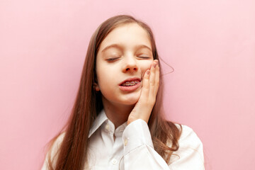 little teenage girl with dental braces suffers from toothache on pink isolated background, child...