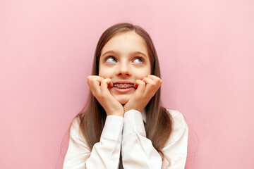 scared teenage girl with braces worries and bites nails on pink isolated background, excited child...