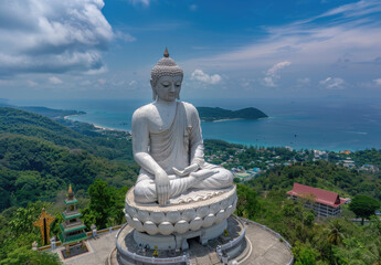 drone view of Phuket Big Buddha, clear blue sky and sea in the background, white statue of sitting buddha on top mountain with green forest around it