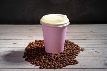 Takeaway coffee coffee cup standing on a roasted coffee beans heap, on a white wood surface with a black textured background