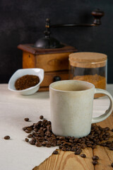 Still life of a cup of coffe sitting in the foreground on a heap of roasted coffee beans. In the background there are an old coffee grinder, a small bowl with grinded coffe and a jar with brown sugar.