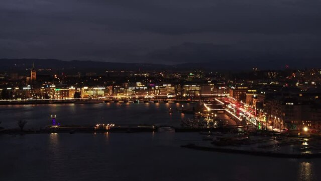 Aerial view of Geneva's waterfront at night, showcasing the city lights and their reflections on the water with Alps in the background