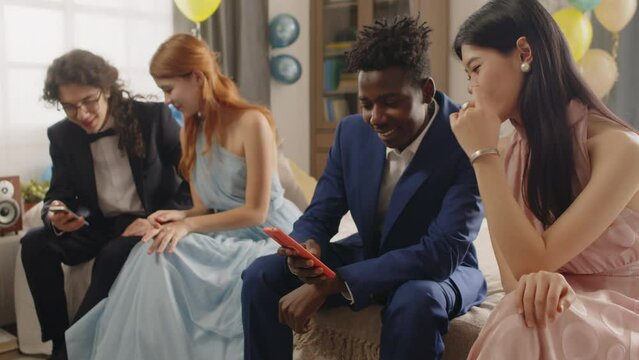 Youthful couple in classy suit and evening gown sitting on bed in domestic room and discussing funny photos from prom party or graduation ceremony against their friends