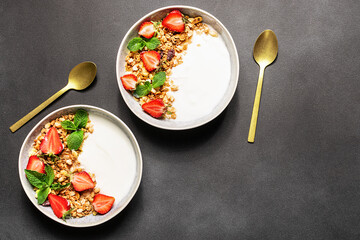 Fresh Greek Yogurt with granola and strawberries decorated with aromatic mint leaves on black stone background top view, copy space for your design. Healthy breakfast concept.
