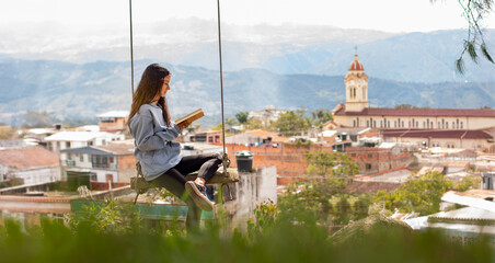 lifestyle: latina woman reads a book sitting on a swing with a beautiful colonial village landscape in the background