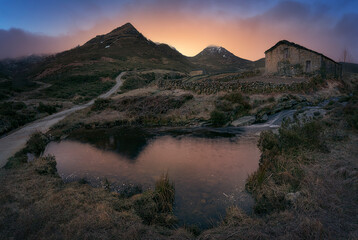 Naklejka premium warm sunrise at the Pardo waterfall, in the mountain pass of Estacas de Trueba, Burgos, with a small pond in the foreground and a pasiega cabin between mountains