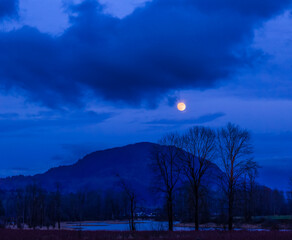 Fototapeta premium A glowing full moon in a cloudy sky over a silhouetted mountain, Village of Harrison Hot Springs; British Columbia, Canada
