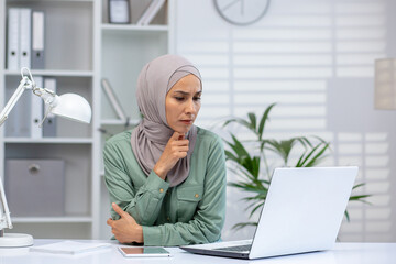 Woman wearing hijab focused while working on a laptop in a well-lit, modern office setting. Professional and thoughtful workspace environment.