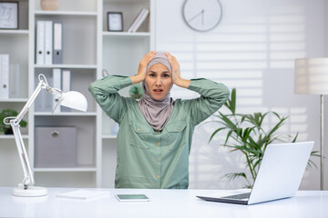 Stressed woman in hijab holding head in frustration while working in modern office. Concept of...