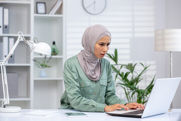 Frustrated woman in a hijab sitting at a desk while working on a laptop in a modern office setting.