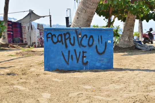 Acapulco, Mexico - April 27 2024: A blue small wall with graffiti on it that says Acapulco Vive after hurricane Otis. The wall is on a sandy beach near a palm tree