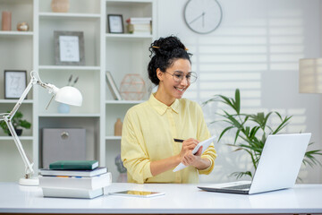 Hispanic woman in a home office environment taking notes and using a laptop, illustrating remote...