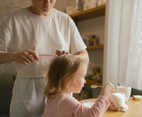 Happy single mother doing her daughter's hair while having breakfast. Woman combs the hair of a...