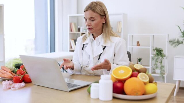 Friendly mature female making video call via wireless device at doctor's workplace in hospital. Attentive dietitian providing online consultation about client's progress in identifying foods.