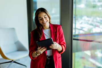 Young professional woman in red blazer using tablet by office window on sunny day