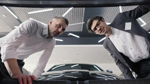 A confident male assistant in a business suit opens the trunk of a car and tells a client a middle-aged man in a white shirt about the features of a particular car model in a car showroom