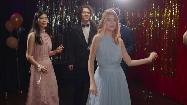 Carefree teenage girl in baby blue evening dress dancing in front of her intercultural friends or classmates at prom party while enjoying spending time with them in the night club