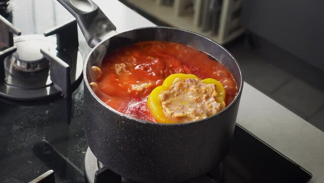 Stuffed peppers with meat and rice cooking in a pot on the stove in red tomato sauce. Sauce is bubbling and steaming, creating a delicious and appetizing food scene. 