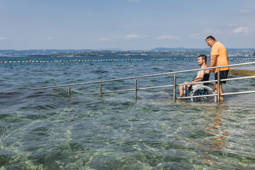 Male assistant helping a man in a wheelchair to enjoy the sea on an access ramp into the water. Accessible beach concept.