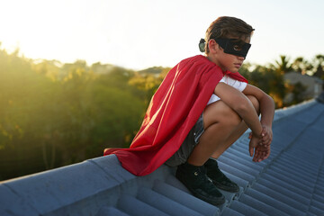 Serious, young boy and playing superhero on roof with costume and mask for fantasy adventure at...