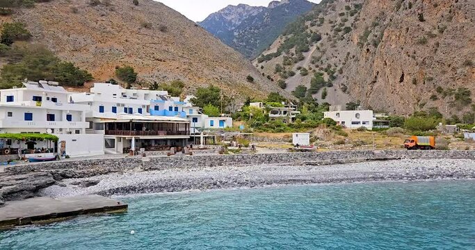 Agia Roumeli village in Crete, with traditional white buildings with blue window shutters, waterfront view with sea, stone beach and mountains in the background. 