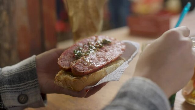 Hombre le pone chimichurri a un choripan, chorizo, pan, chimi. Comida, asado, s&aacute;ndwich, parrillada.