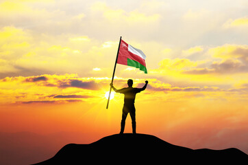 Oman flag being waved by a man celebrating success at the top of a mountain against sunset or sunrise. Oman flag for Independence Day.