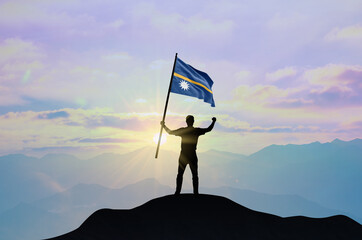 Nauru flag being waved by a man celebrating success at the top of a mountain against sunset or sunrise. Nauru flag for Independence Day.