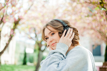Beautiful young woman in comfortable clothes listens to music in headphones in blooming park. Sakura Pink flowers. Spring.