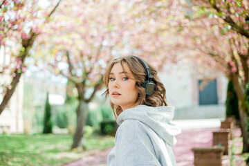 Beautiful young woman in comfortable clothes listens to music in headphones in blooming park....