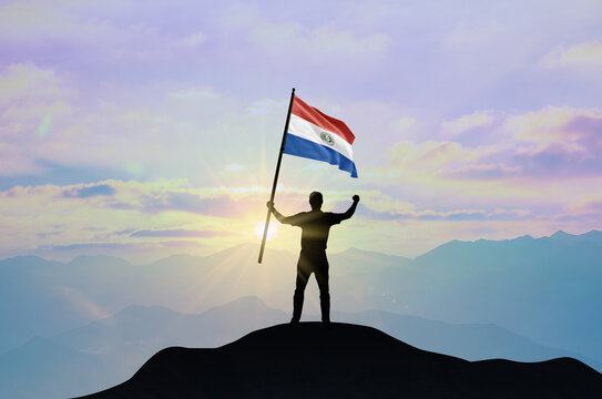 Paraguay flag being waved by a man celebrating success at the top of a mountain against sunset or sunrise. Paraguay flag for Independence Day.