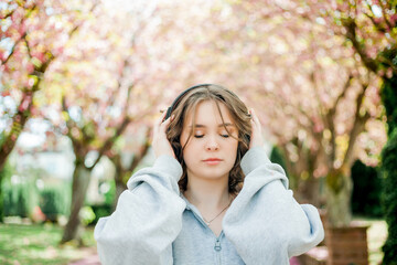 Beautiful young woman in comfortable clothes listens to music in headphones in blooming park....