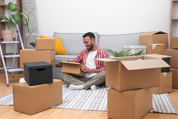 Young male with beard sits in new house unpacking things brought by courier in large cardboard...