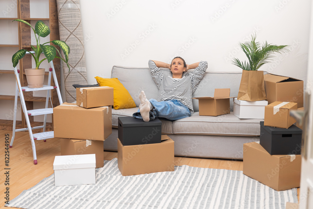 Poster woman sits on soft sofa waiting for help of husband in collecting boxes with furniture standing near
