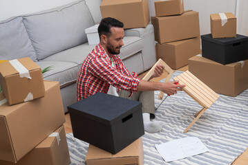 Smilling man sits with wooden furniture among various carton boxes with stuff for interior...