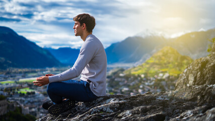 A young man peacefully meditates at sunset in a rock overlooking the mountains and the sea. A Caucasian guy does yoga in simple asanas on a viewpoint in the sunset sunlight