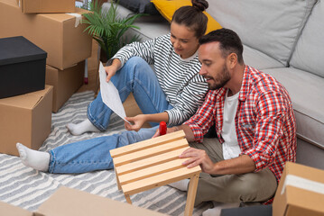 Young couple, amidst boxes in their new home, happily assembles modern wooden shelf furniture,...