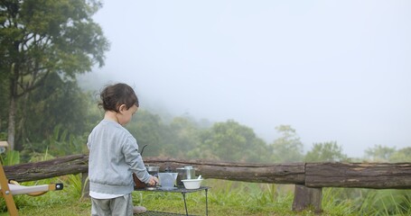 Cute Asian child girl is standing at a picnic table with a cup in her hand to dipping coffee in the...