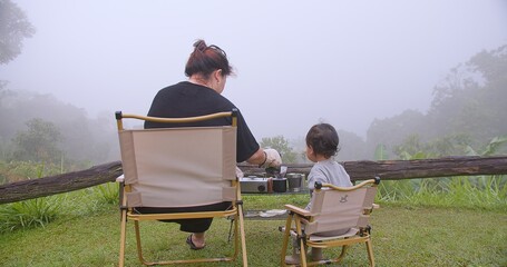 A woman and a child are sitting in chairs outside, enjoying a meal together. The atmosphere is peaceful and intimate, with the two of them sharing a moment bonding over food at campsite camping tent