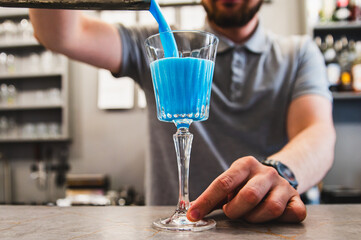 A person pours blue liquid into a clear stemmed glass with a blue straw on a bar counter.