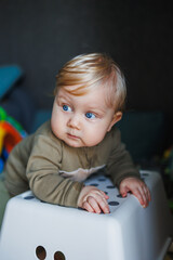 Fototapeta premium A small baby boy with blue eyes and blond hair is standing near a chair. A cute emotional baby is learning to stand on his feet