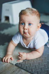 A small child in a white bodysuit is learning to crawl on a clean, heated floor. Home photo of a cute blue-eyed baby.