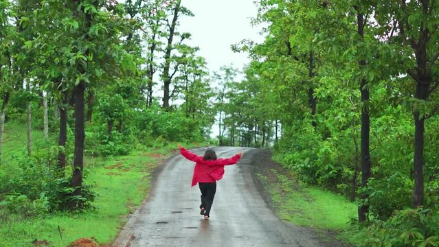 Happy Indian teenager girl running on the road covered with green trees on both side during monsoon season at Saputara, Gujarat, India. Girl running with raised arms in forest during holidays. 