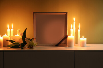 Blank funeral frame, burning candles and lily flowers on cabinet in dark room