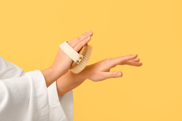 Young woman massaging her arm with body massage brush on yellow background