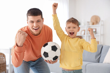 Young man and his little son cheering for football team with soccer ball in living room