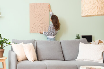 Young woman hanging picture on turquoise wall in stylish living room