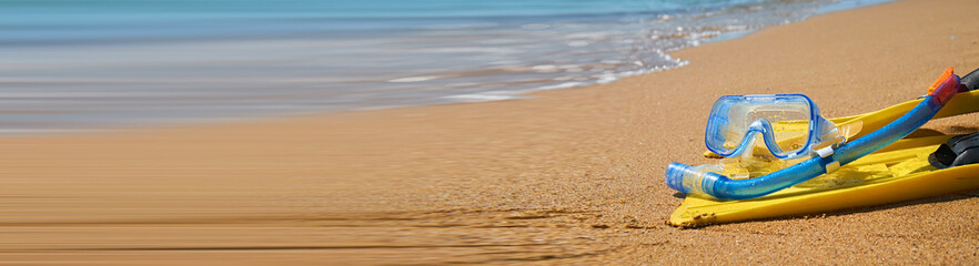 Banner of bright yellow flippers and a blue snorkel lie on the soft sand of a tropical beach. The turquoise ocean waves gently lap at the shore in the background. © Pihuliak